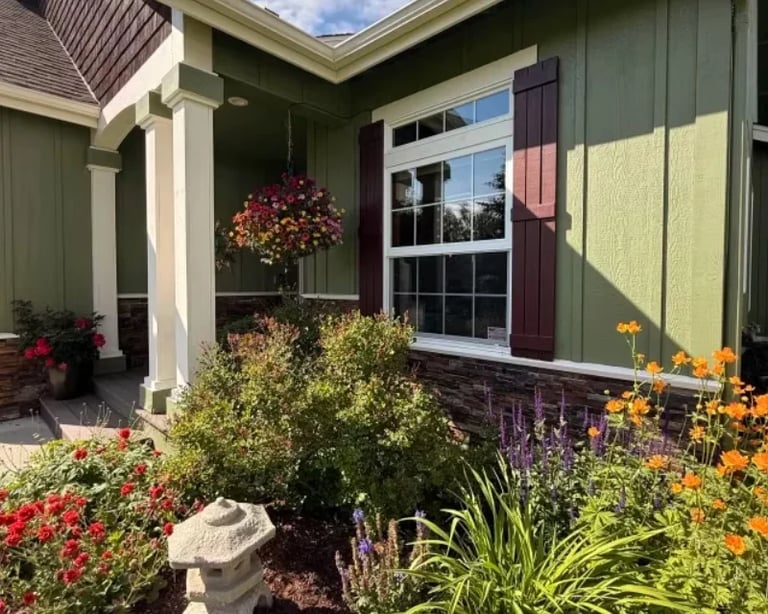 Modern home entrance with green exterior, burgundy shutters, covered porch, and colorful flower garden featuring red, yellow, and purple blooms
